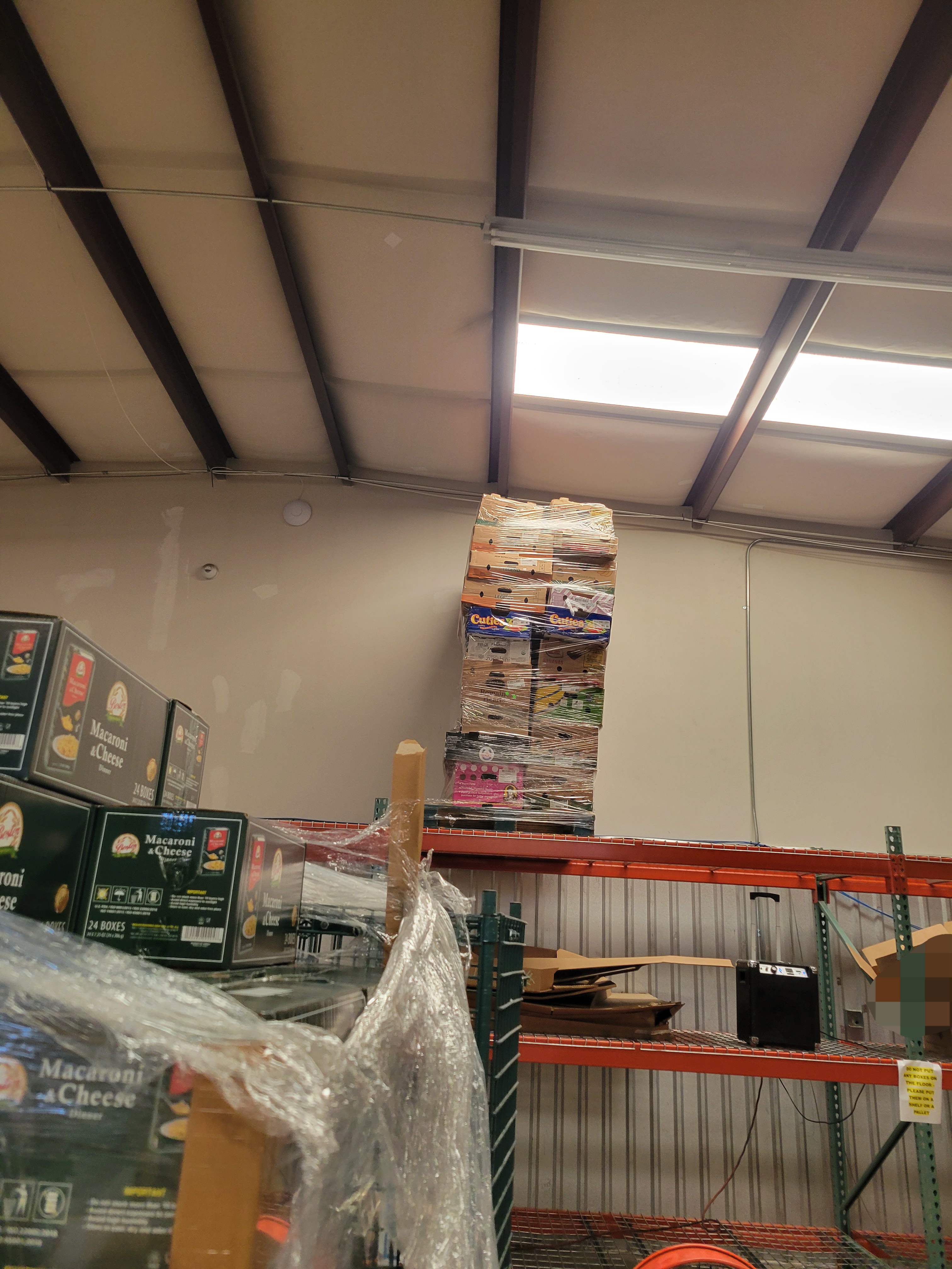 Banana and fruit boxes wrapped in plastic wrap on a pallet on the top shelf with a pallet of boxes of mac n cheese in frame.